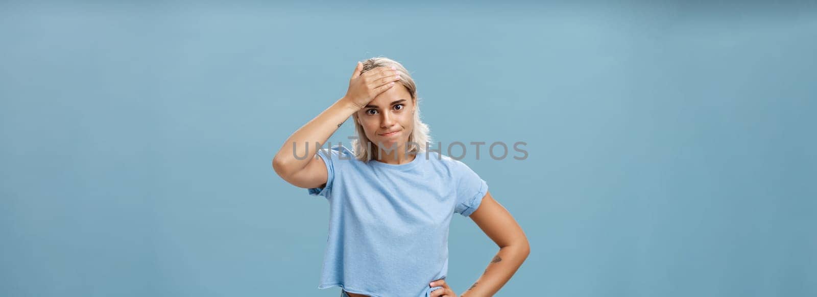 Lifestyle. Girl cannot deal with naughty kids being left alone while mom on shopping holding hand on forehead breathing out losing temper being in troublesome perplexed situation tired over blue background.