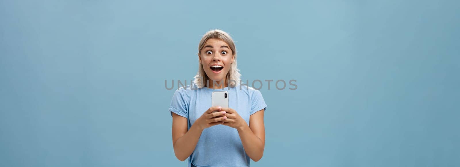 Lifestyle. Indoor shot of amazed attractive blonde female coworker in casual t-shirt gasping from amazement and happiness being surprised staring surprised at camera holding smartphone receiving awesome news.