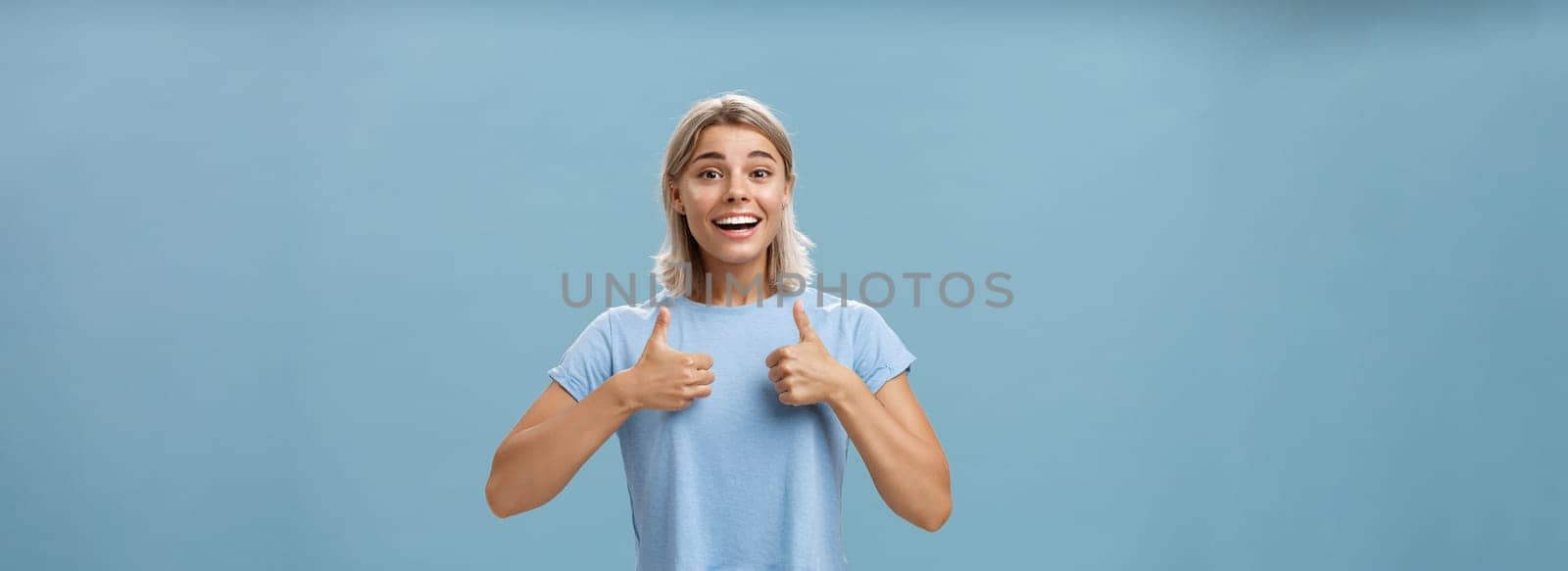 You did great proud of you. Portrait of satisfied and fascinated attractive happy girlfriend in casual t-shirt showing thumbs up and smiling broadly being supportive and cheerful over blue wall.
