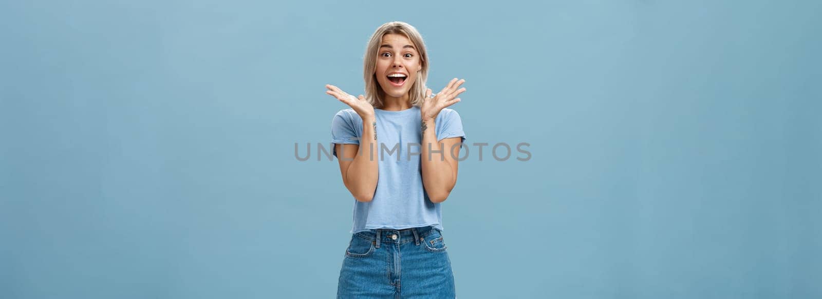 Thrilled enthusiastic happy woman in trendy t-shirt and shorts clapping hands from amazement smiling broadly and gazing with admiration at camera being surprised and joyful over blue wall. Copy space