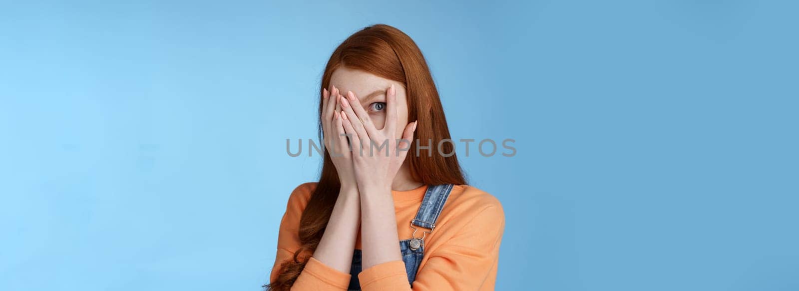 Not peeking promise. Charming intrigued cute redhead teenage girl hiding face cover eyes palms look through fingers check out gift anticipating something interesting standing curious blue background.