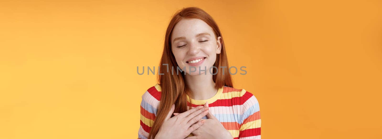 Happy romantic delighted sighing young lovely redhead girlfriend smiling broadly touching heart chest palms close eyes recalling date passionate kiss feel gratitude warmth, orange background by Benzoix