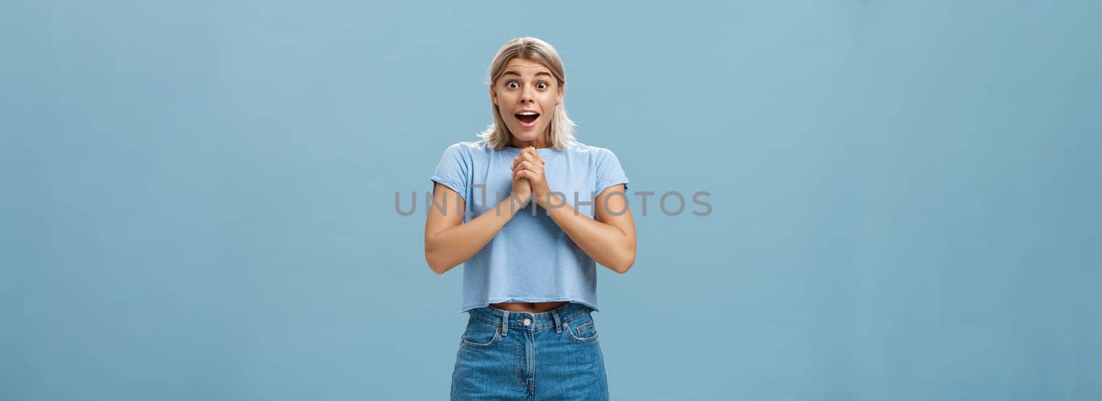 Lifestyle. Surprised girl receiving bouquet of flowers from secret fan holding hands together on breast gasping opening mouth from amazement and happiness popping eyes from astonishment over blue wall.