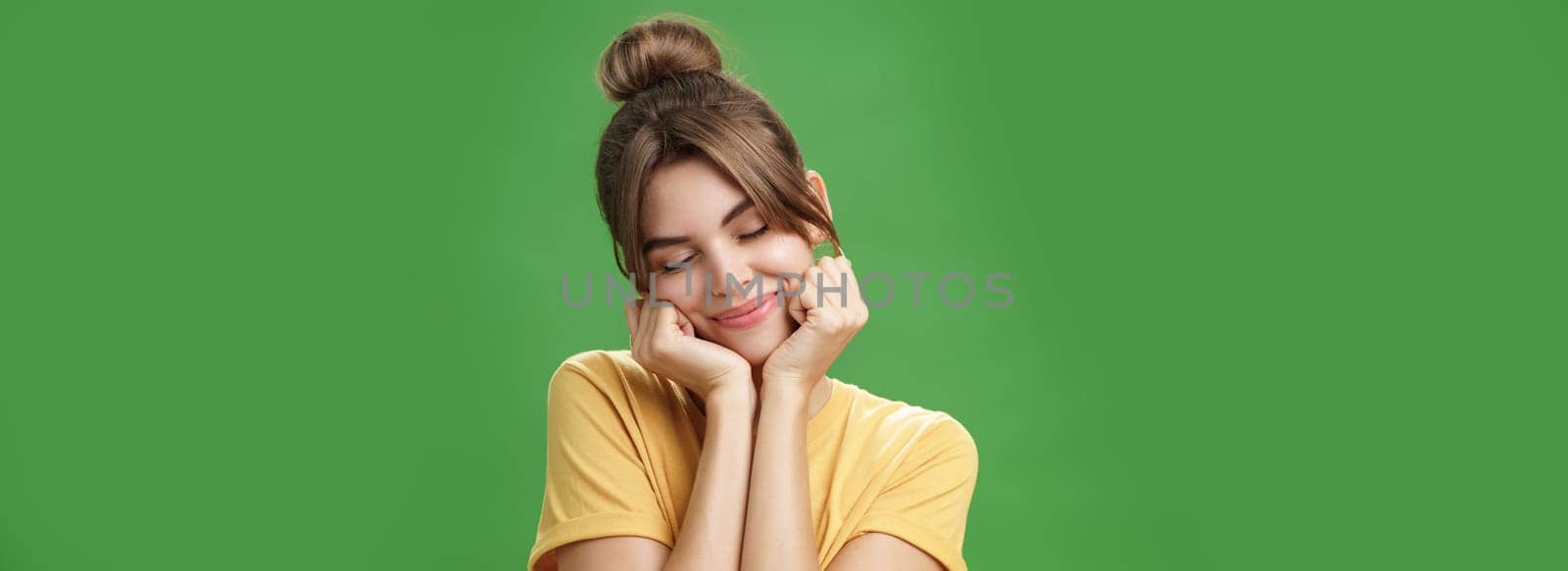 Tender sensual timid european girl in yellow t-shirt leaning head on shoulder touching cheeks with hands closing eyes and smiling with soft grin feeling nostalgic and romantic over green background. Lifestyle.