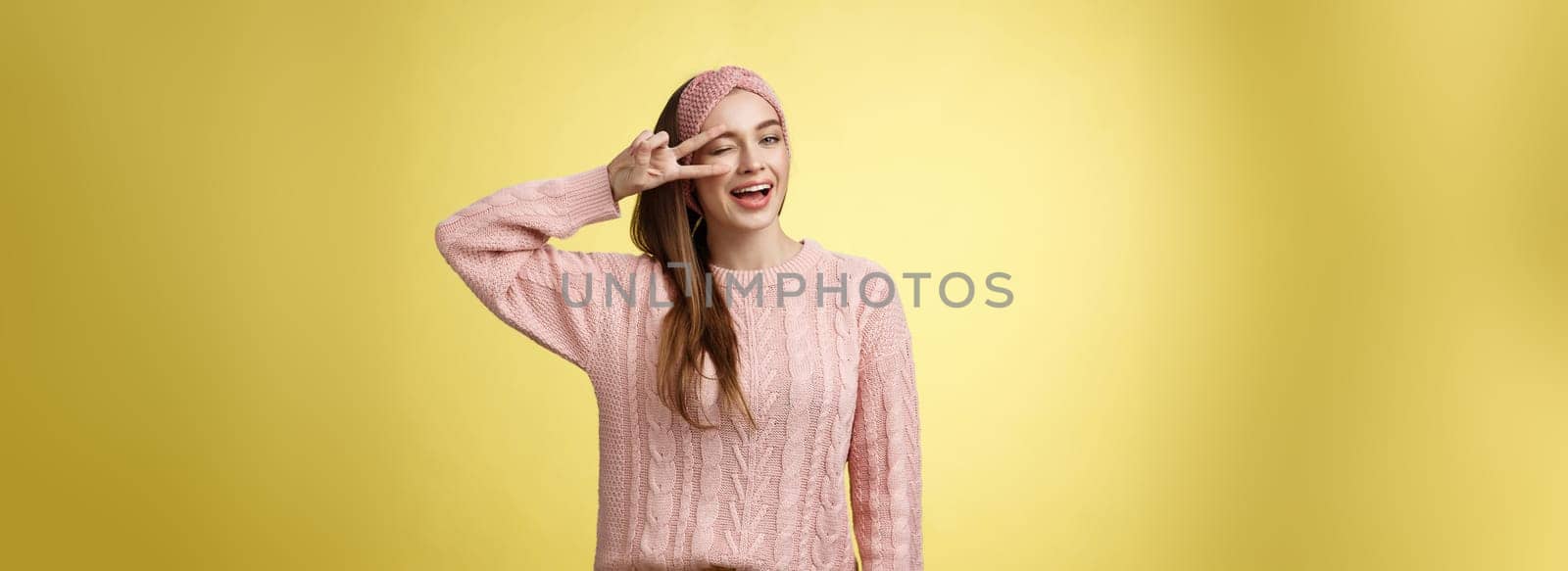 Cheerful happy glamour young european woman in pink knitted sweater, wearing headband, winking flirty and cute, showing victory or pease sign over eye, feeling excited and joyful over yellow wall.