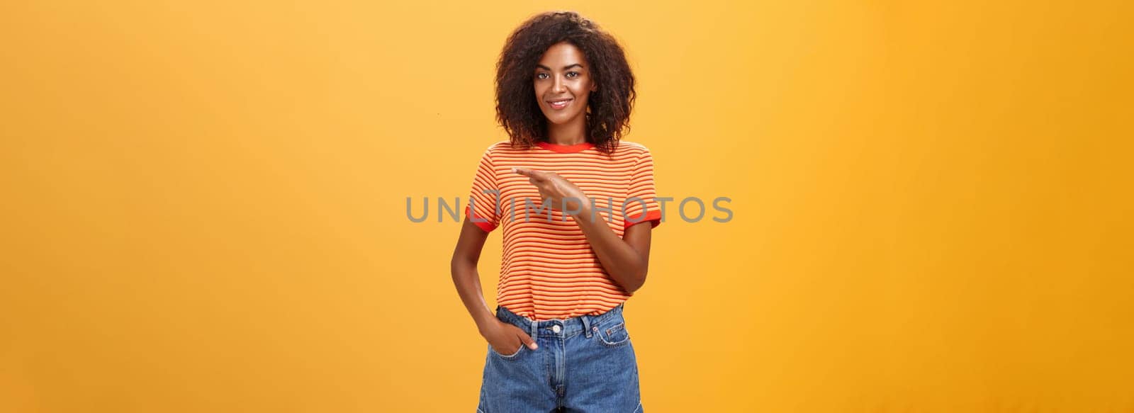 Showing perfect place for advertising. Charming carefree and confident young stylish african-american woman with afro hairstyle holding hand in pocket casually pointing right over orange wall.