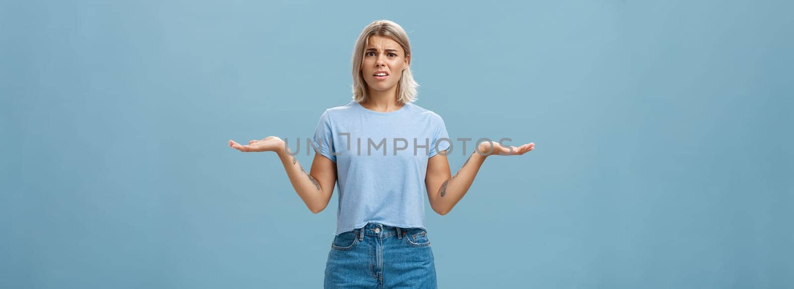 Girl feeling struggles to understand what happening. Confused perplexed intense good-looking female with fair hair shrugging with hands spread aside in displeased clueless pose posing over blue wall.