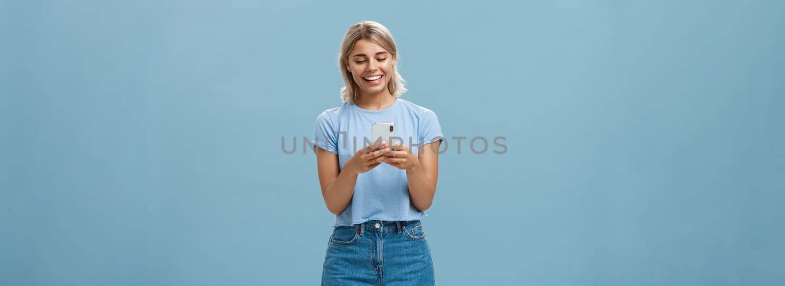 Nice to have internet friends. Carefree happy attractive blonde female student with tanned skin and blond hair reading message from smartphone smiling at device screen pleased over blue wall by Benzoix