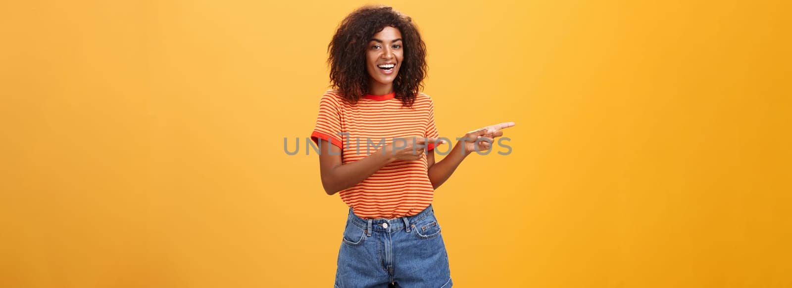 Ask him. Portrait of friendly and joyful good-looking stylish female shop assistant with curly hair and dark skin pointing left with both hands, smiling assured and entertained over orange background by Benzoix