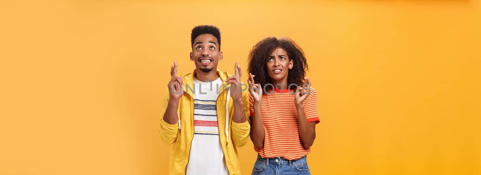 Couple of friends praying and looking up in sky intense crossing fingers for good luck clenching teeth faithfully hoping parents not saw them smoking standing together over orange background by Benzoix