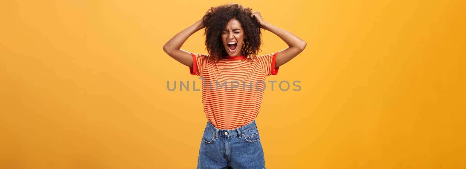 I hate life being cruel. Depressed african woman in rage and fury pulling hair out of head yelling and screaming with closed eyes losing temper having bad day hating everything over orange wall by Benzoix