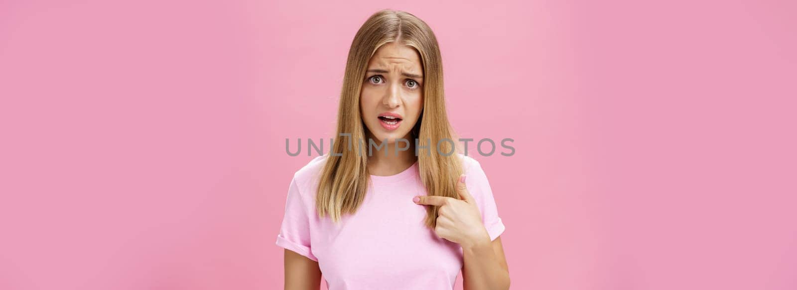 Insulted girl pointing at herself with displeased pissed and questioned expression asking question being shocked she picked or accused in something terrible and disrespectful posing against pink wall. Lifestyle.
