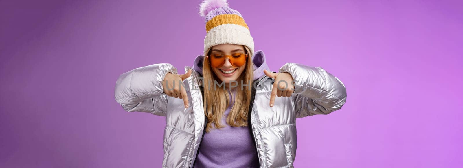 Joyful stylish blond woman in silver jacket stylish sunglasses jat enjoying perfect mountains view snowy vacation resort look pointing down smiling amused having fun feel happy, purple background.