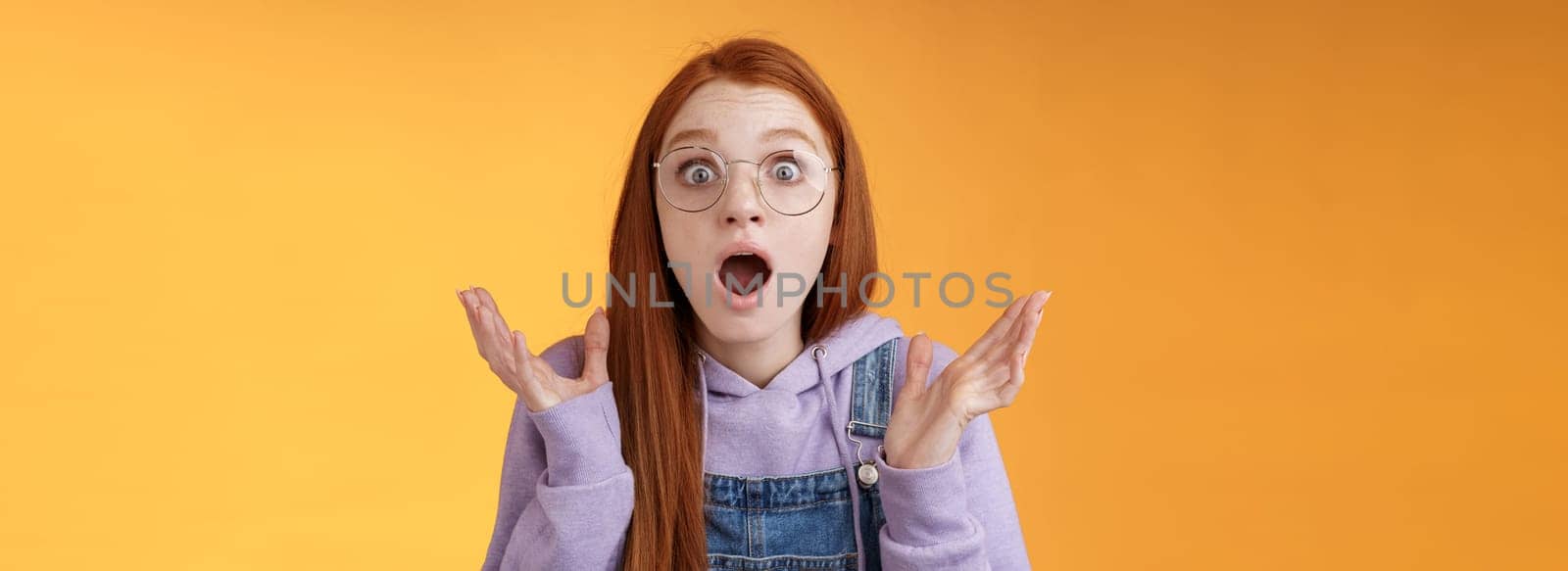 Close-up shocked sensitive concerned young panicking redhead woman worry drop jaw gasping raise hands spread freak out stare surprised emotional reacting incredible news, orange background.