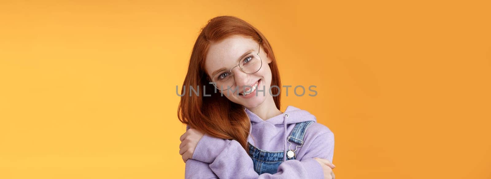Passion, tenderness, wellbeing concept. Girl accept own self smiling charming grin tilt head hugging herself embracing body feel happiness delighted relaxing, flirty gaze camera orange background.