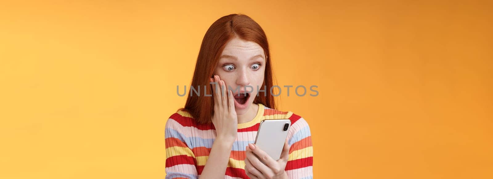 Omg what heck. Portrait shocked concerned young redhead sensitive impressed redhead woman stare smartphone display touch cheek drop jaw stunned surprised standing orange background hold phone.