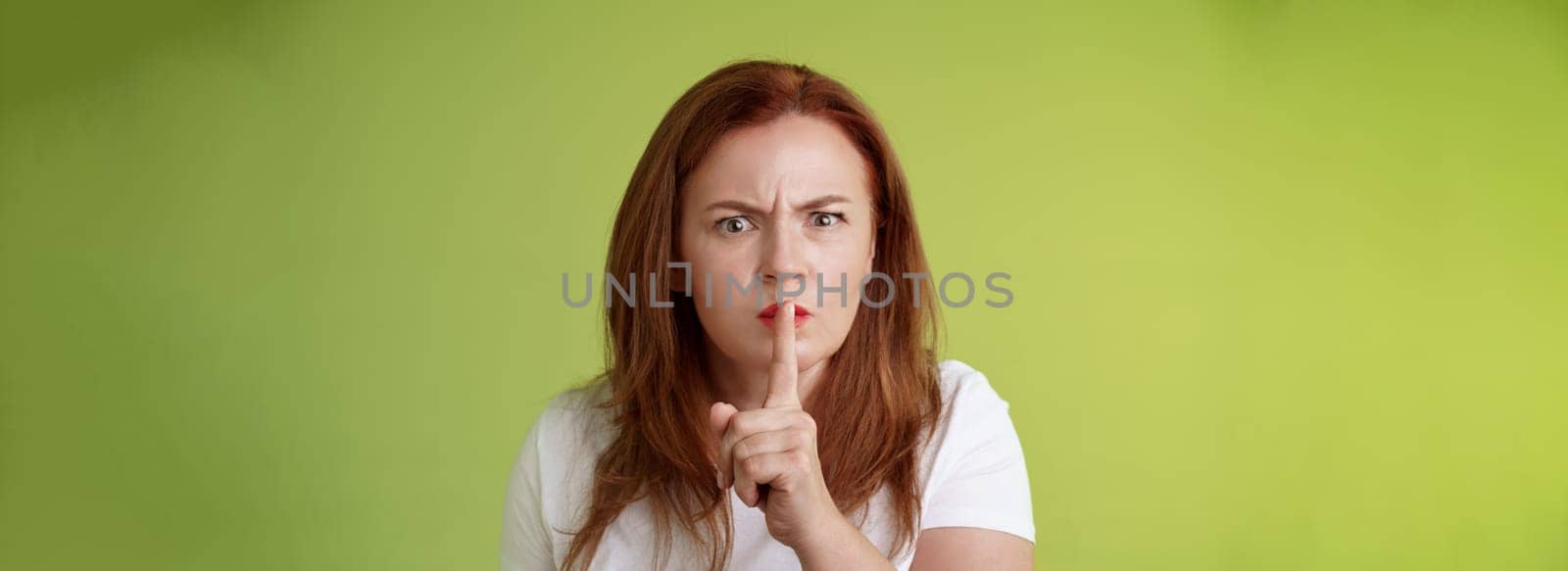 Not speak during exam. Strict serious-looking displeased middle-aged redhead woman frowning disappointed hushing say shush index finger pressed lips keep quiet gesture green background by Benzoix