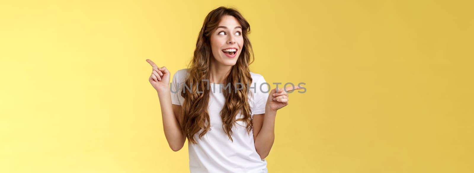Amused cheerful smiling happy young curly-haired woman chestnut hair observe curiously copy space grinning admiration joy pointing sideways making choice impressed satisfied turn left.