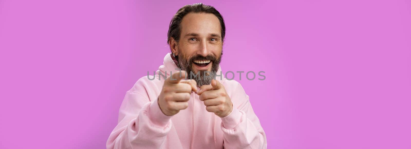 Funny amused carefree happy adult handsome man pranking friend fool around pointing camera index fingers greeting choosing you laughing joyfully having fu, standing purple background rejoicing.