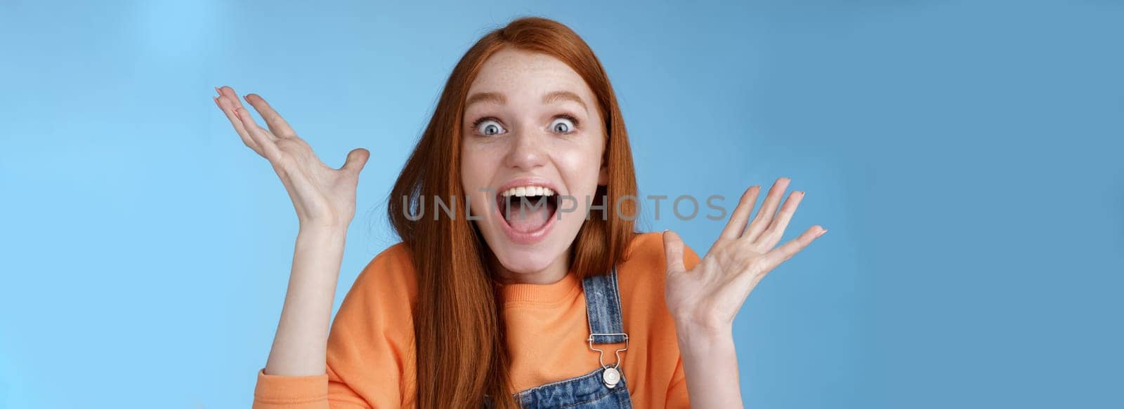 Surprised astonished sensitive overwhelmed young happy redhead girl receive incredible fantastic prize wide eyes astonished raising hands triumphing win lottery celebrating joyfully.