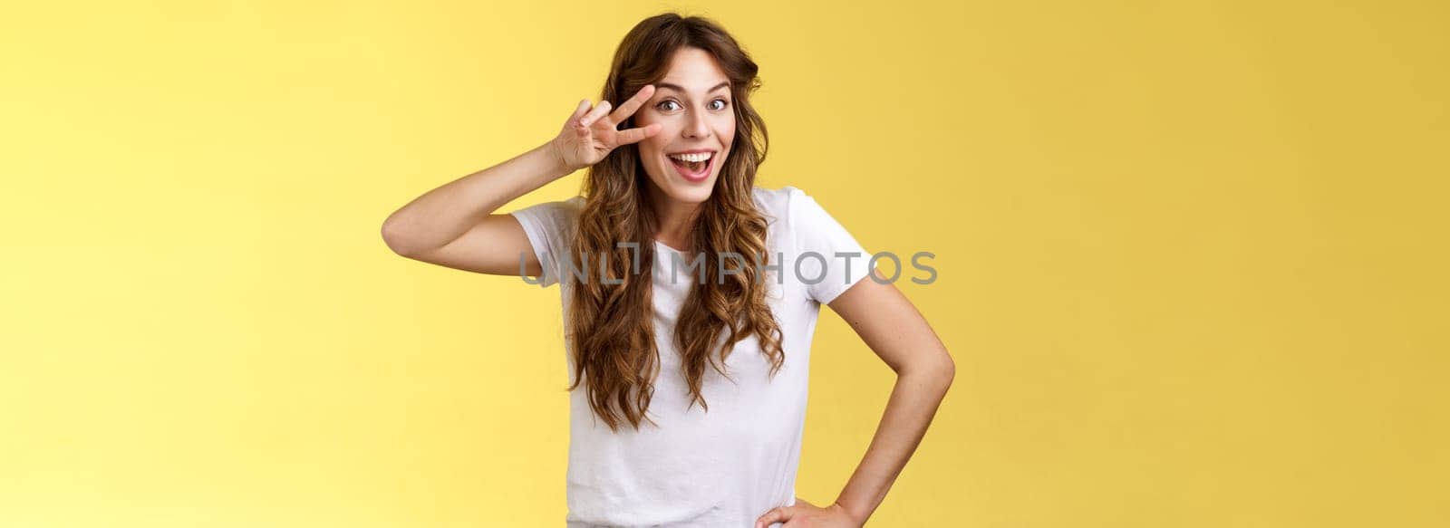 Playful surprised charismatic young caucasian girlfriend having fun look amused enthusiastic show peace victory sign laughing smiling joyfully enjoy summer holidays touch hip stand yellow background. Lifestyle.