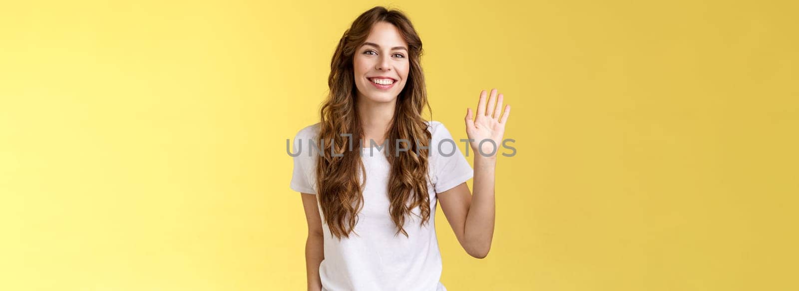 Outgoing comfortable relaxed friendly casual european woman curly haircut waving raised palm say hello introduce herself become new member smiling broadly greeting hi gesture yellow background. Lifestyle.