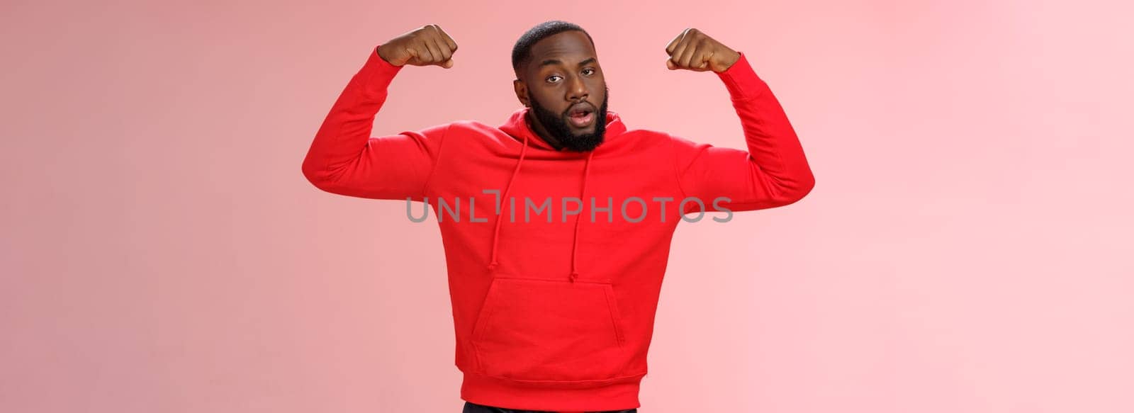 Strong cheeky serious-looking african american sportsman in red hoodie bragging showing-off raise hands excercising show big muscles biceps, standing confident pink background by Benzoix