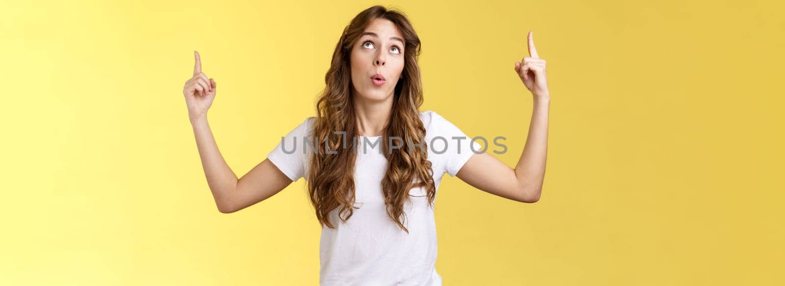 Impressed curious wondered attractive european woman long curly haircut look pointing index fingers up top copy space promo react astonished fascinated surprising event yellow background.