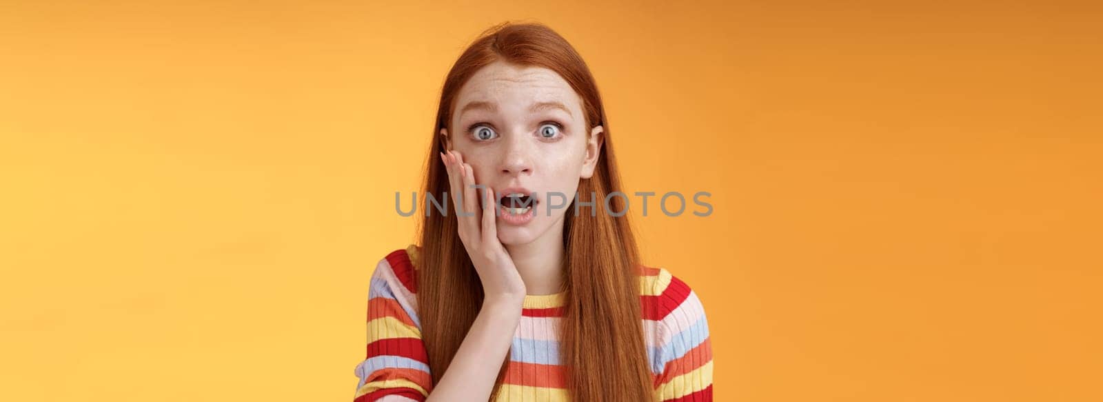 Concerned worried ginger girl blue eyes drop jaw gasping touch cheek confused looking nervously anxious, show empathy hearing terrible upsetting story standing orange background. Copy space