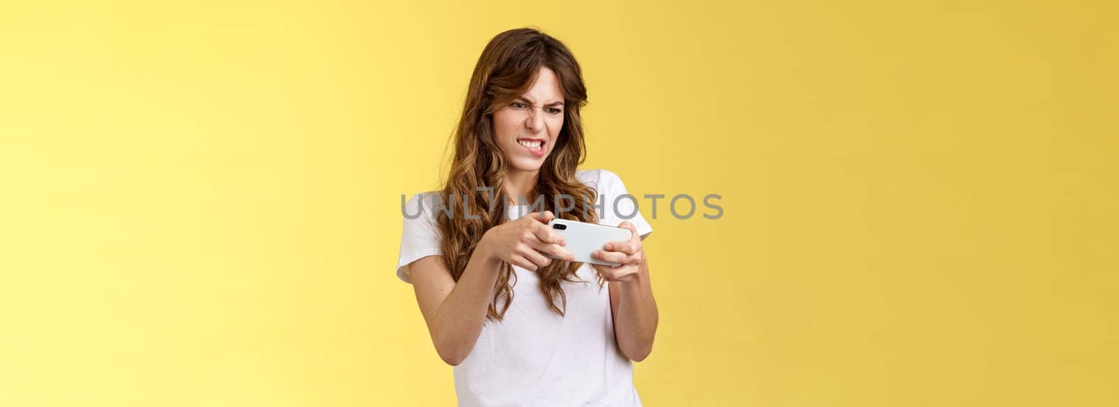 Excited focused playful enthusiastic geeky attractive girl trying beat score hold smartphone horizontal grimacing intense look screen playing awesome arcade game yellow background.