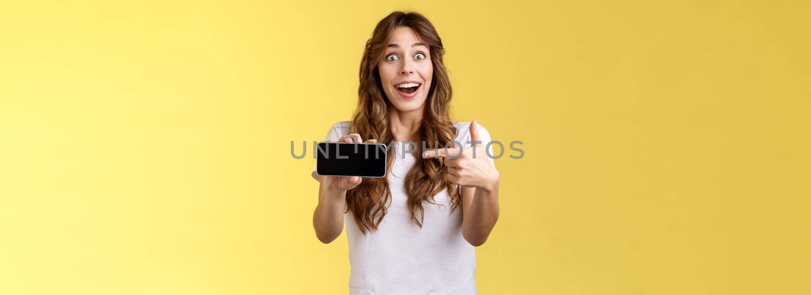 Impressed upbeat happy lucky girl curly long hairstyle open mouth admiration joy like awesome new app show smartphone screen horizontal phone display stand yellow background amazed by Benzoix