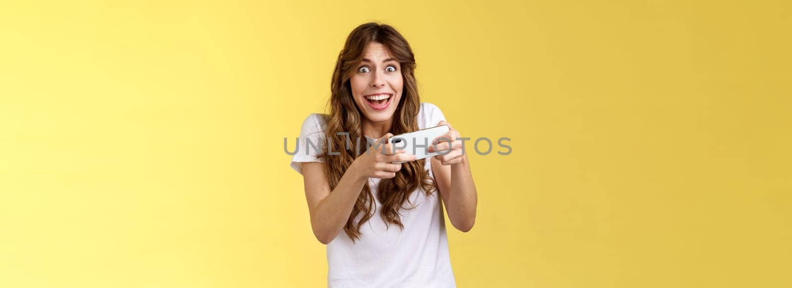 Happy enthusiastic excited cute playful girl winning awesome smartphone game thrilled playing stare camera surprised triumphing smiling happily hold mobile phone horizontal yellow background.