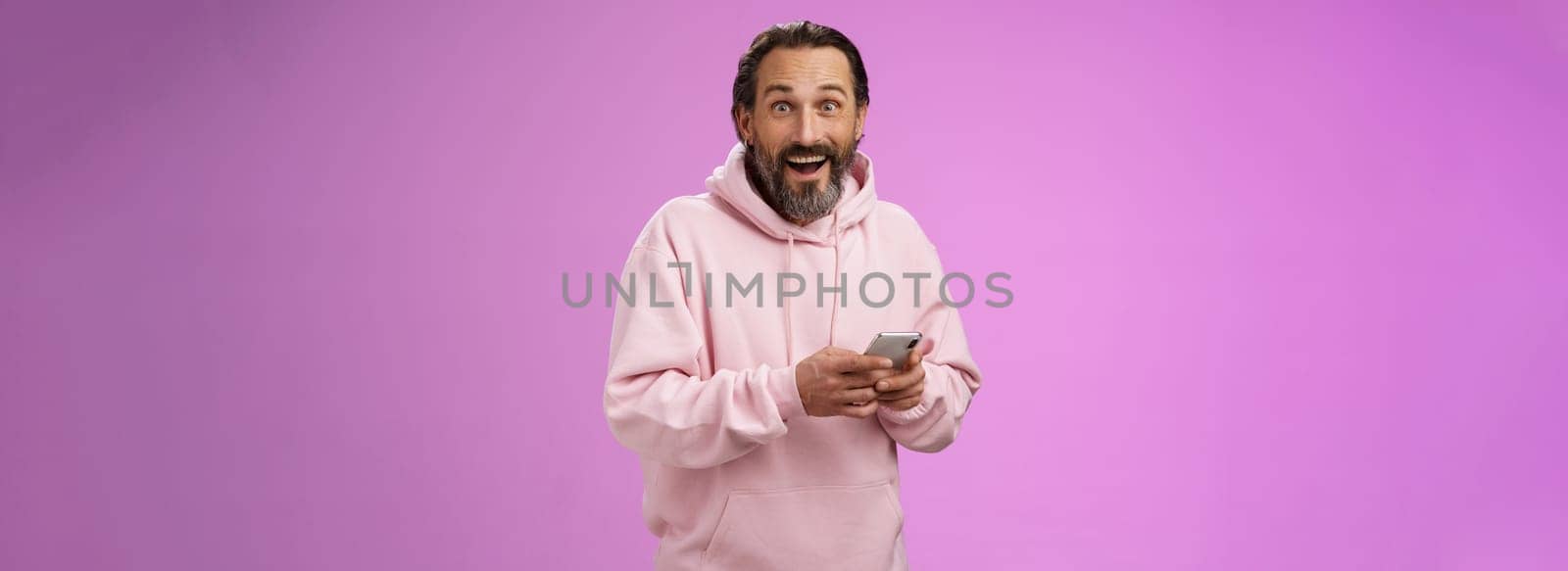 Amused happy fascinated adult mature caucasian bearded man in pink hoodie gasping impressed holding smartphone look camera astonished purchase cool new phone, astonished how convenient.