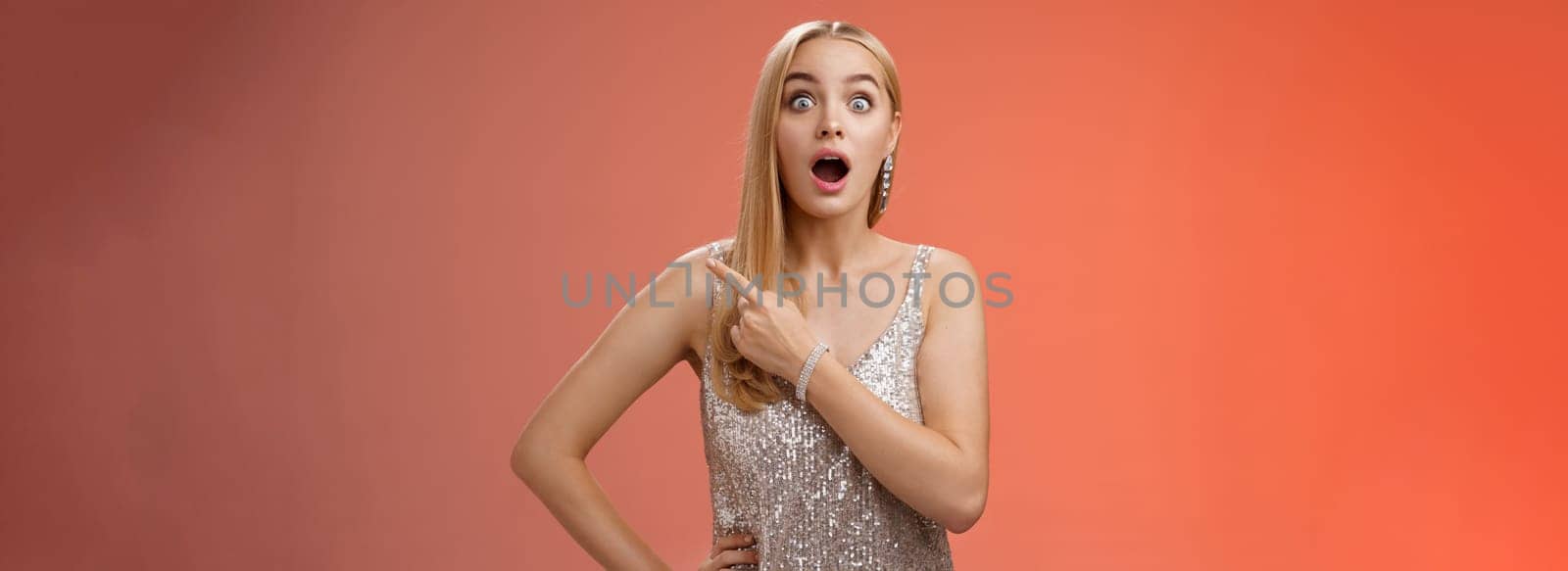 Amused speechless impressed european blong girl drop jaw widen eyes thrilled pointing upper right corner excited amazed standing red background gossiping wealthy bartender wear silver shiny dress by Benzoix
