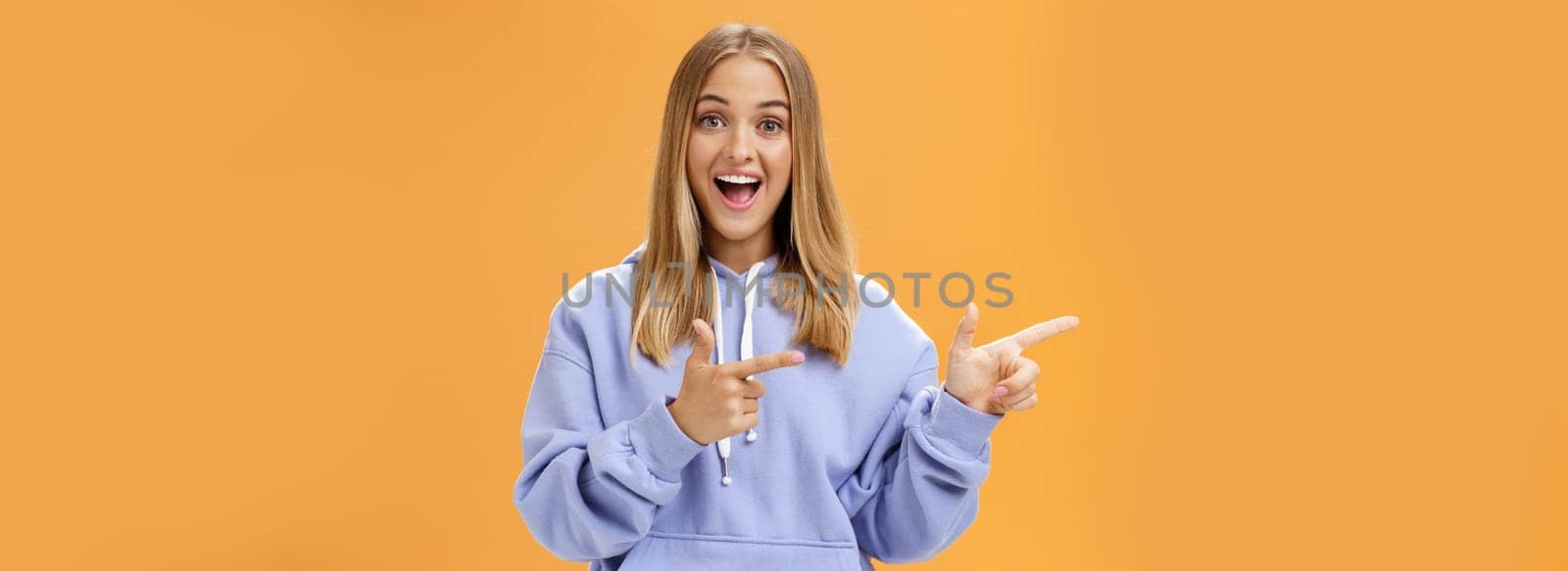 Amused girl pointing at curious copy space showing left with index fingers smiling excited and surprised standing entertained with upbeat grin in cozy blue hoodie over orange background. Lifestyle.