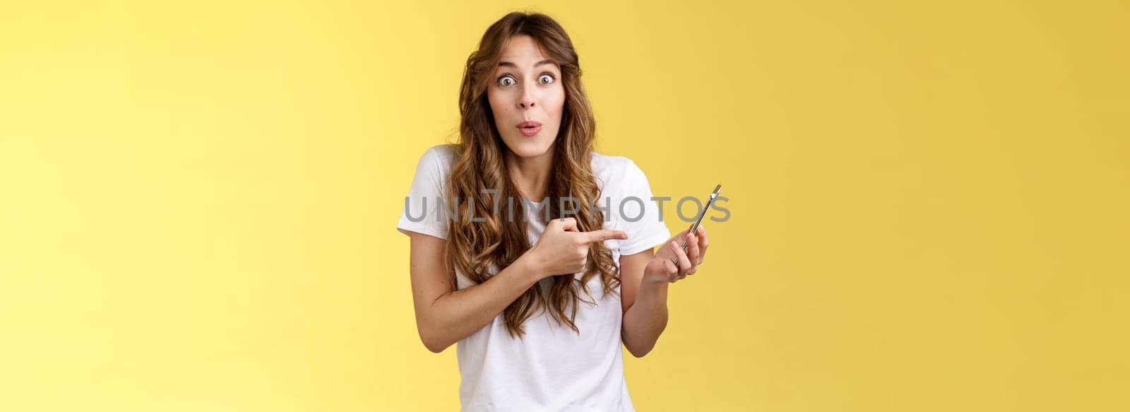 Impressed enthusiastic good-looking girl curly cute hairstyle folding lips amused wow stare camera astonished hold smartphone pointing mobile phone speak about awesome news yellow background by Benzoix