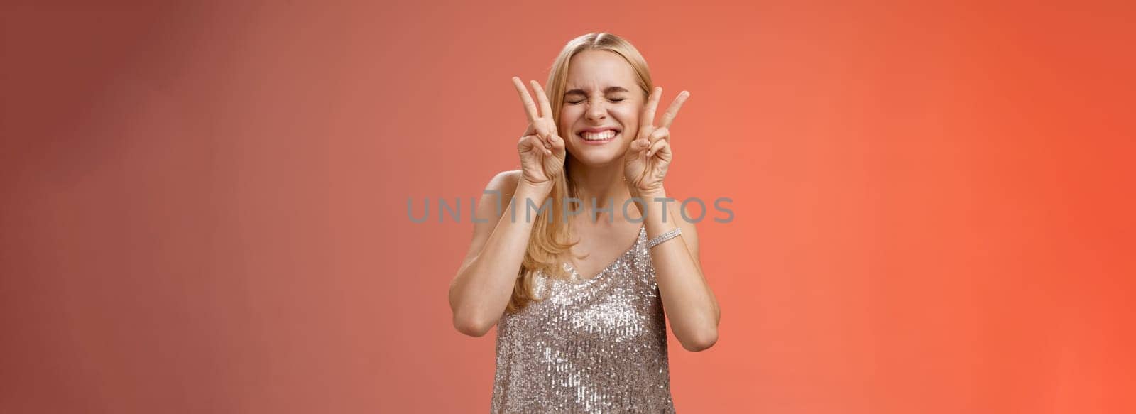 Excited carefree happy cheerful european blond woman have fun drink cocktails dancing partying nightclub girlfriend smiling closed eyes show peace bunny gesture red background. Copy space