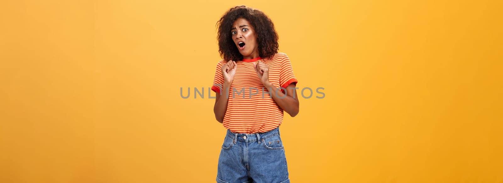 No do not hurt me. Scared silly and insecure good-looking african american woman in stupor screaming for help turning away raising palms near body in defence standing in fright over orange wall by Benzoix