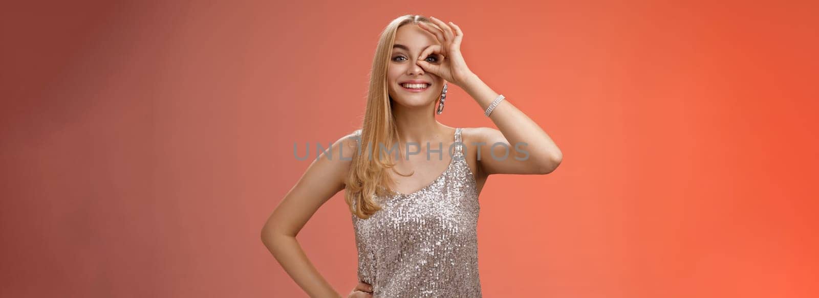Charming elegant blond european carefree woman in silver glittering evening dress show no problem okay sign look through eye smiling delighted check out awesome promo intrigued, red background.
