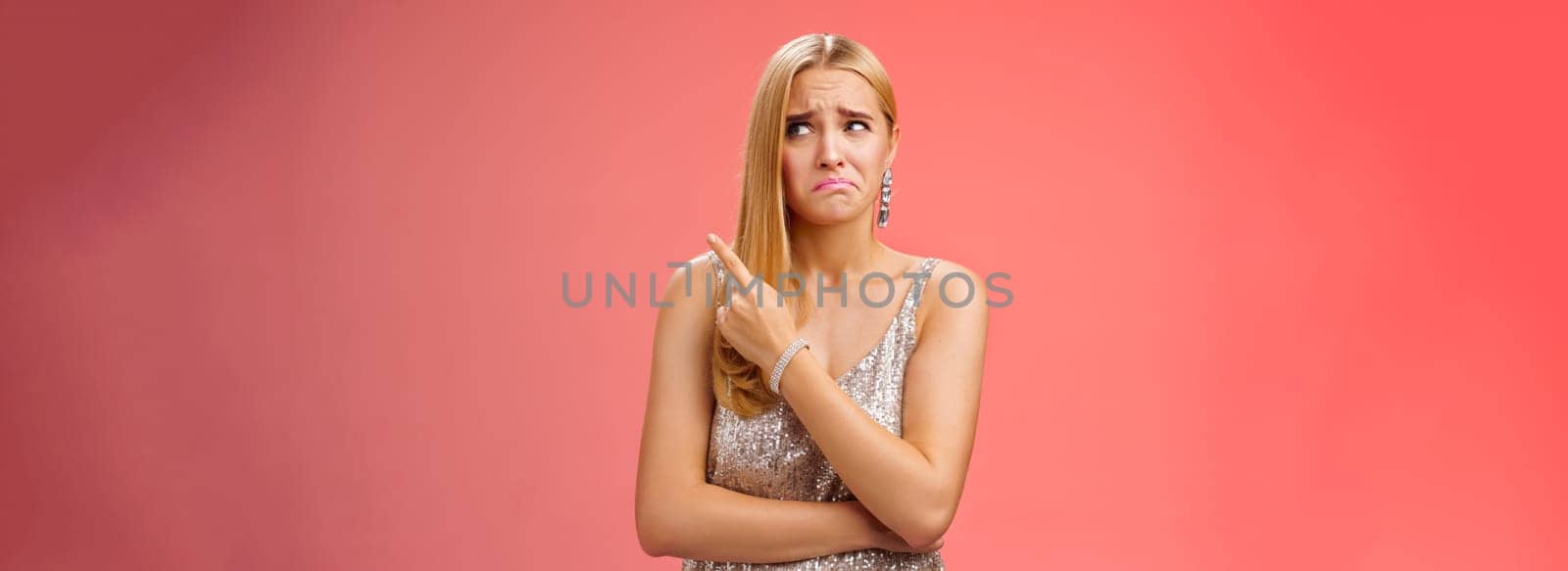 Upset doubtful unsure cute blond funny glamour girl in silver evening dress cringing grimacing hesitant pointing looking upper right corner suspicious, uncertain standing red background by Benzoix