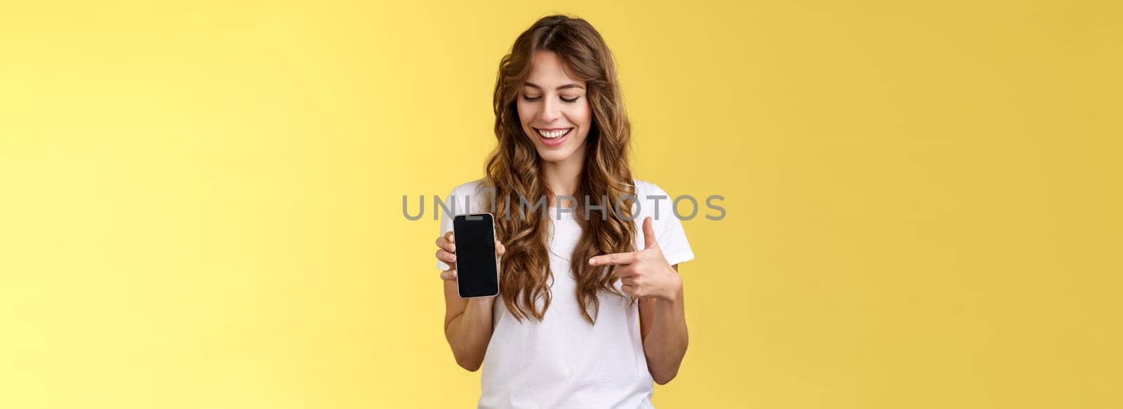 Intrigued charismatic happy young female blogger show what photo app use edit social media profile hold smartphone looking pointing index finger mobile phone blank screen smiling amused by Benzoix