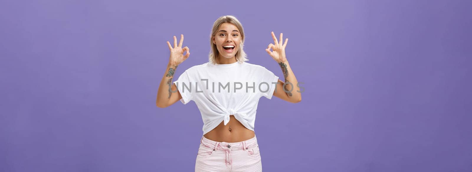 Girl being agree with friends deciding were enjoy holidays. Portrait of charming satisfied and happy young blonde female with tattoos and pierced belly showing okay gesture and smiling with delight. Copy space