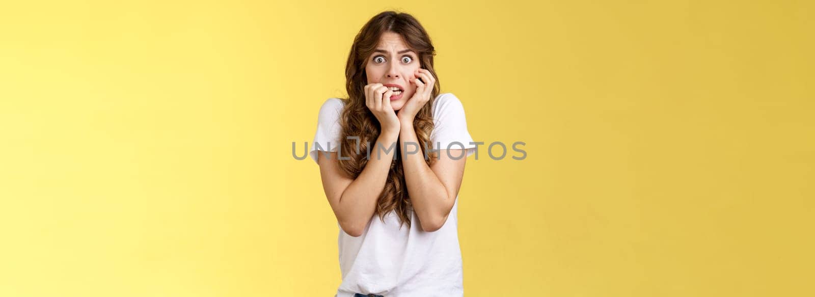 Girl panicking feeling scared timid insecure biting fingernails stare camera frightened clench teeth trembling fear touch face nervously feeling unconfident terrified stand yellow background.