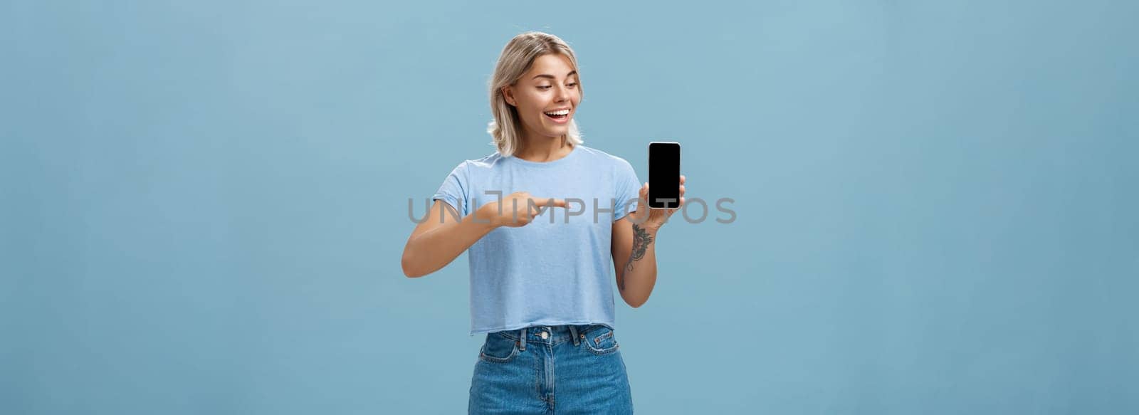 Portrait of delighted good-looking european female with blond haircut showing smartphone screen with amazement and delight pointing at device while promoting brand new gadget over blue background. Technology concept
