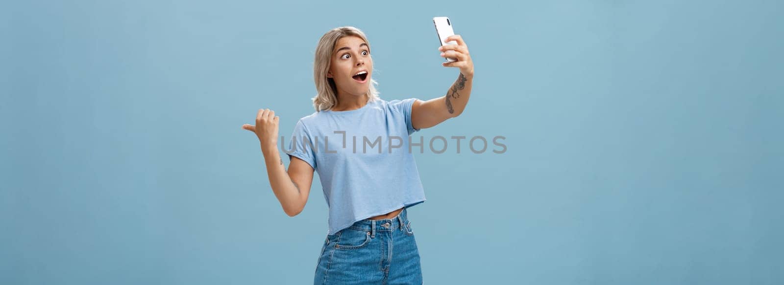 Lifestyle. Girl recording video showing followers awesome scene attending cool event holding smartphone looking impressed and surprised at device screen pointing backwards indicating right over blue wall.