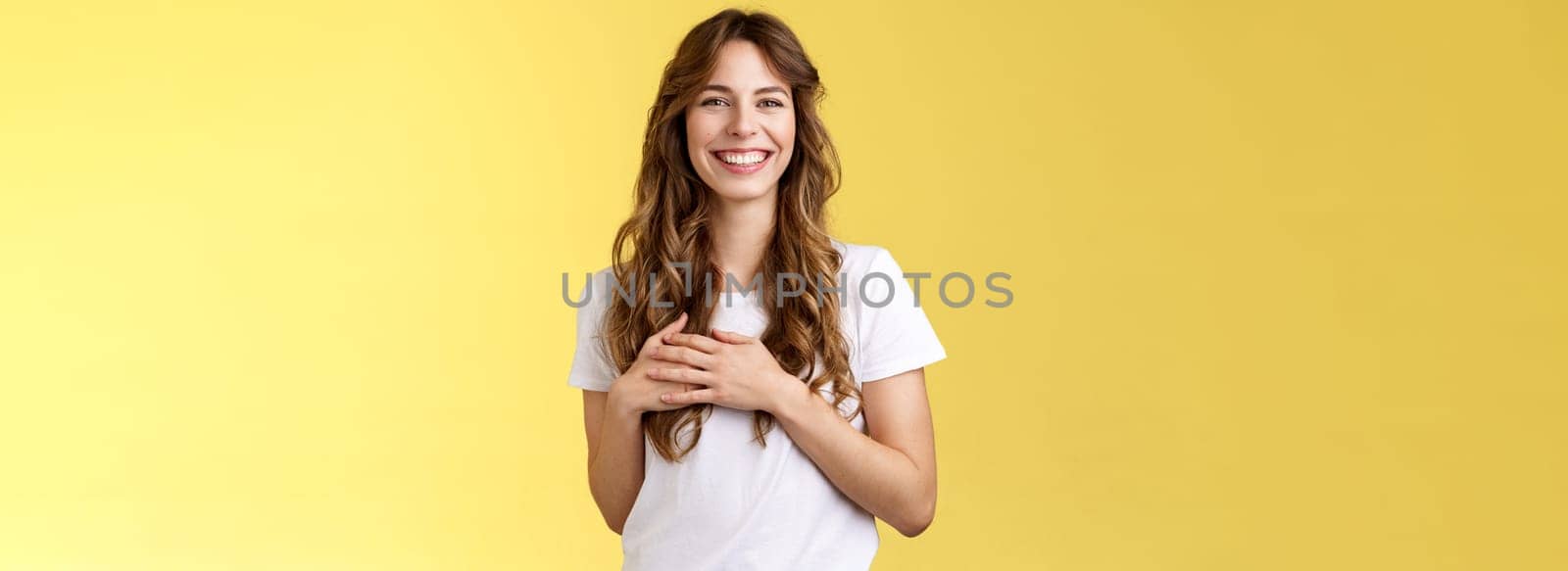 Tender heartwarming kind gentle feminine girl curly long hairstyle look sincere delighted squinting joyfully smiling broadly touch heart hold hands chest grateful lovely gaze stand yellow background. Lifestyle.