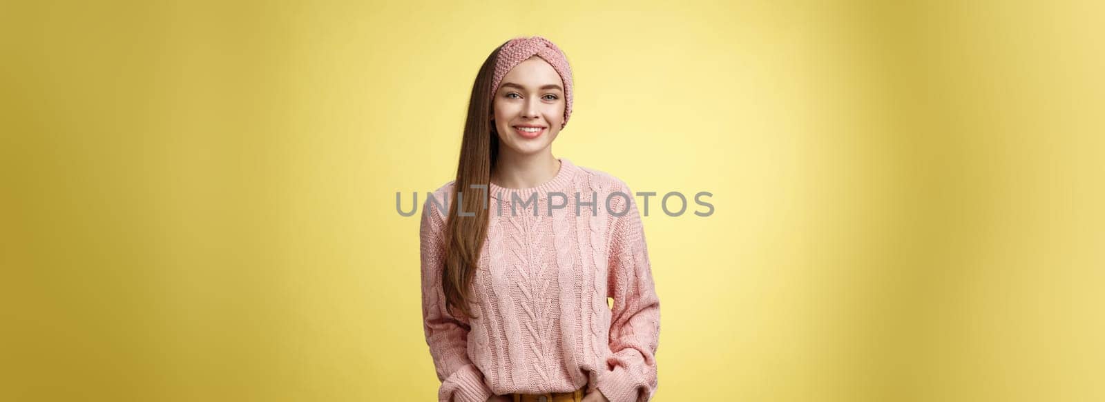 Charming friendly young girl in knitted sweater and headband holding hands in pockets, smiling, looking positive motivated achieving goals having great day, posing positive against yellow background by Benzoix
