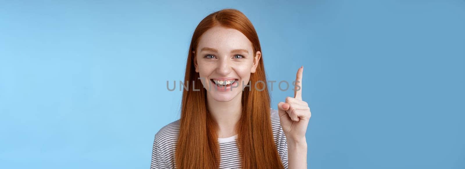 Got excellent idea pick for you. Attractive happy friendly-looking helpful redhead female shop assistant showing gorgeous dress customer pointing up index finger smiling glad help, blue background.