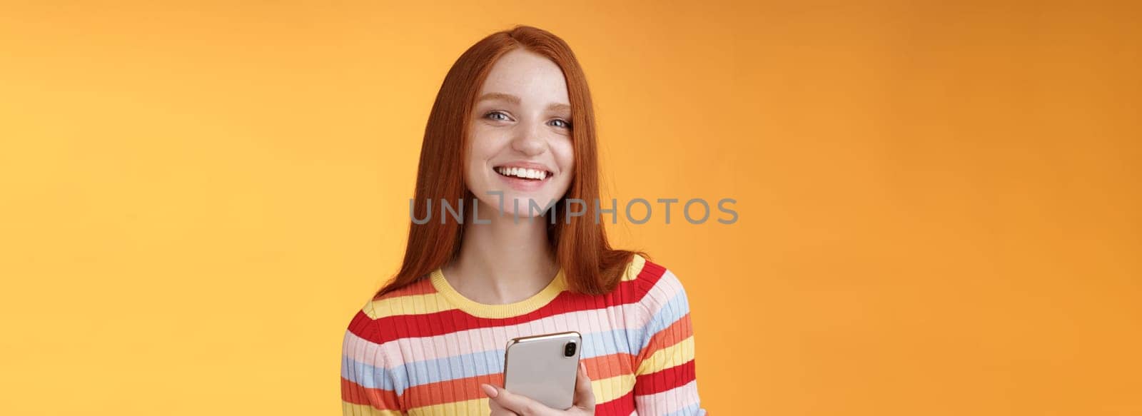 Friendly enthusiastic young redhead girl blue eyes using smartphone turn camera answer smiling broadly telling who sent message standing delighted orange background messaging, texting boyfriend by Benzoix