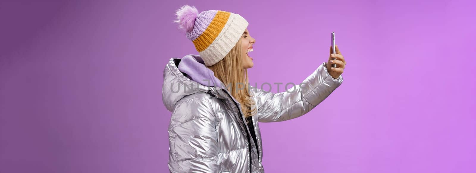 Amused carefree attractive caucasian blond girl in silver winter jacket hat extend arm holding smartphone recording own yell open mouth wide close eyes fool around mimicking funny faces selfie by Benzoix